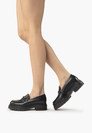 Mocassins noirs en cuir avec une semelle épaisse, dotés d'une boucle décorative et d'une texture lisse. Vue latérale mettant en avant la forme et le design.