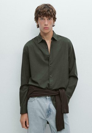 Dark green button-up shirt with a sleek design, paired with light blue jeans and a brown, ribbed sweater wrapped around the waist.