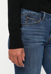 Ranka šalia klubo, vilkinti tamsiai mėlynus džinsus su užtrauktuku priekyje ir juodą ilgomis rankovėmis marškinėlį su grioveliais ant rankovių.