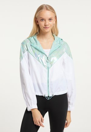 Giovane donna che indossa una giacca bianca con cappuccio e spalle azzurro chiaro lucido e leggings neri, in piedi davanti a uno sfondo semplice.