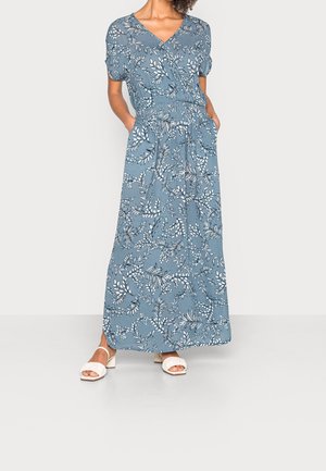 Kvinde iført en blå blomsterprint slå-om kjole med korte ærmer og lommer, kombineret med hvide åbne hælede sandaler.