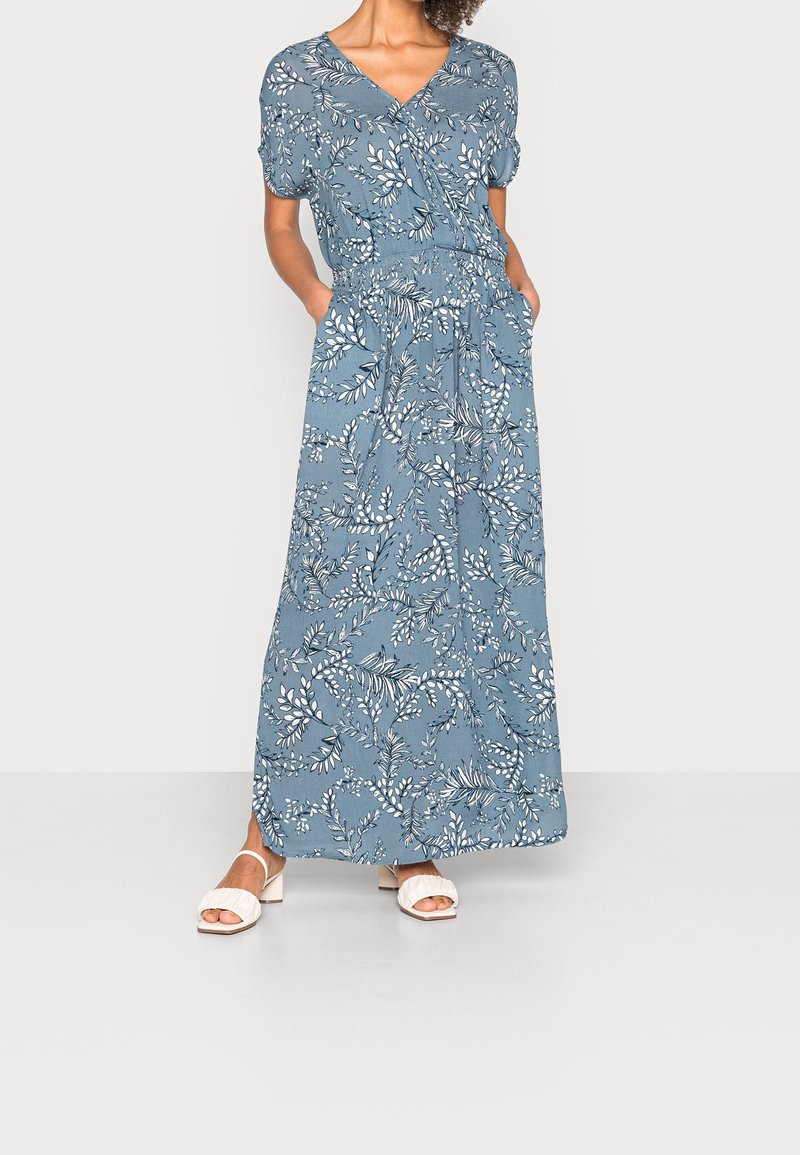 Femme portant une robe portefeuille bleue à fleurs avec manches courtes et poches, associée à des sandales blanches à talons et à bouts ouverts.