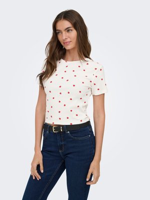 Mujer con camisa blanca de manga corta con pequeños patrones de rosas rojas y vaqueros azul oscuro con cinturón negro, de pie contra un fondo claro.