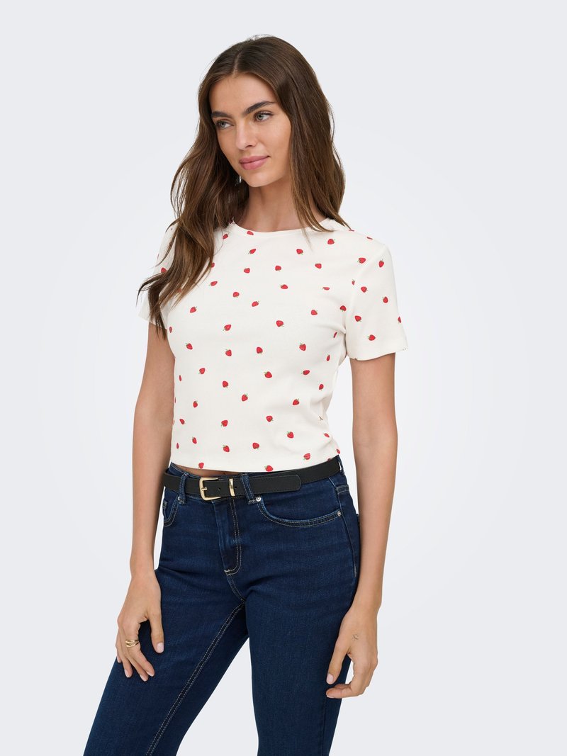 Femme portant une chemise blanche à manches courtes avec de petits motifs de roses rouges et un jean bleu foncé avec une ceinture noire, debout devant un fond clair.
