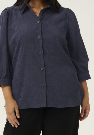 Kobieta w granatowej bluzce zapinanej na guziki z bufiastymi rękawami do połowy przedramienia i czarnych spodniach na jednolitym tle.