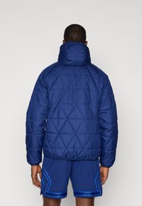 Vojna prešita puffer jakna v temno modri barvi z visokim ovratnikom in elastičnim robom. Zasnovana za toplotno zaščito, ima gladko teksturo in diamantni vzorec.