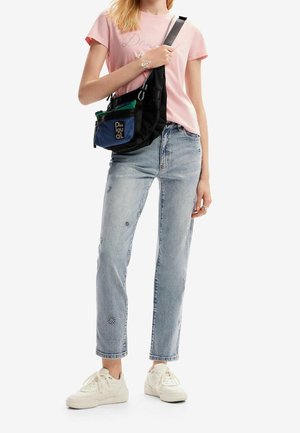 Kvinde iført lyserød t-shirt, lyseblå broderede jeans, hvide sneakers og en sort crossbody taske med en blå frontlomme.