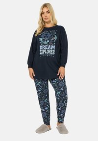 Granatowy zestaw piżamy składający się z długiego rękawa z napisem "Dream Explorer" oraz kosmicznymi grafikami, w połączeniu z wzorzystymi legginsami i szarymi kapciami.