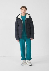 Navy pufferjack met een crèmekleurige fleece-voering in de capuchon, gecombineerd met een teal trui en bijpassende sweatpants. Draagt lichte sneakers.