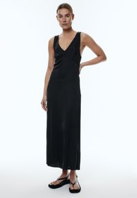 Zwarte mouwloze maxi-jurk met V-hals, een gladde textuur en een getailleerde taille, gecombineerd met zwarte sandalen met bandjes en een plateauzool.