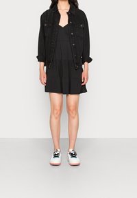 Czarna dżinsowa kurtka, bezrękawnikowa czarna sukienka z falbanami oraz białe sneakersy z czarnymi sznurowadłami i różowymi podeszwami, stojąca na neutralnym tle.