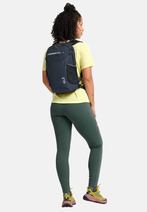 Femme aux cheveux bouclés portant un sac à dos bleu marine, un T-shirt jaune, des leggings verts et des chaussures de randonnée jaunes, debout de profil à droite sur un fond blanc.