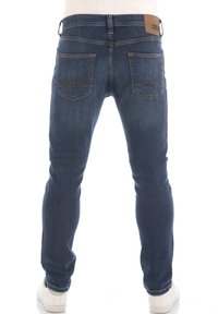 Jeans de denim azul de corte ajustado con una textura sutil, cinco bolsillos y detalles de costura en contraste. Parche de logo de cuero marrón en la cinturilla.