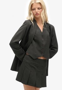 Sort stribet blazer og tætsiddende vest lagd over en kort plisseret nederdel, der har knapdetaljer, struktureret form og glat overflade.
