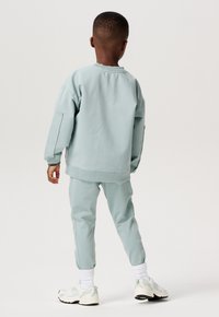 Helle grüne Sweatshirt und passende Jogginghosen aus weichem Stoff. Das Sweatshirt hat überschnittene Schultern; die Jogginghosen haben Bündchenabschlüsse. Weiße Turnschuhe.