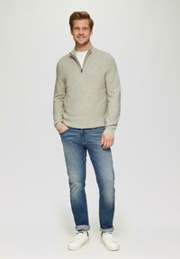Hellgrauer strukturierter Pullover mit Reißverschluss und Rautenmuster, kombiniert mit blauen Jeans und weißen Sneakers. Das Model steht lässig mit den Händen in den Taschen.
