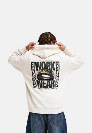 Osoba z warkoczami odwrócona tyłem, zakłada białą bluzę z kapturem z napisem "WORK WEAR" i grafiką opony na plecach, ubrana w niebieskie dżinsy z logo DC.