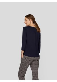 Marineblaues Langarmshirt mit einer Faltendetail auf der Rückseite, kombiniert mit gemusterten Hosen, die geometrische Designs in Marineblau und Beige aufweisen.
