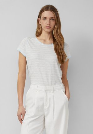 Donna con lunghi capelli castano chiaro, che indossa una t-shirt azzurro chiaro a maniche corte e pantaloni bianchi a vita alta, in piedi con una mano in tasca.