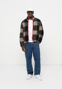 Mikina z flísu s šedým, čiernym a béžovým kockovaným dizajnom, červeným lemovaním, oblečená na biele tričko a kombinovaná s modrými džínovými nohavicami a bielymi teniskami.