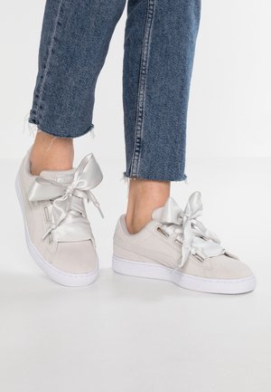 Osoba nosząca jasnobeżowe sneakersy z szerokimi sznurowadłami z satynowej wstążki i przycięte niebieskie jeansy na białym tle.