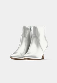 Paari läikivaid hõbedasi pahkluu saapaid, millel on teravad varbad, külgvahe ja läikivad kõrged kontsad, lihtsal valgel taustal.