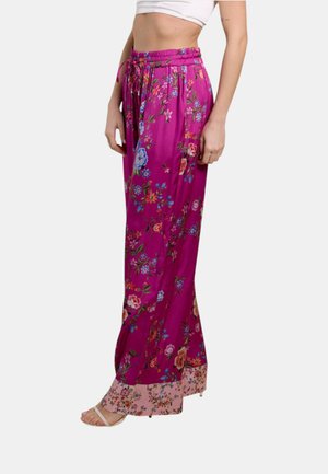 Donna che indossa pantaloni magenta a vita alta e gambe larghe con motivi floreali e cintura annodata, abbinati a sandali bianchi con punta aperta.