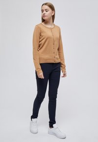 Cardigan marrone chiaro con scollatura rotonda e bottoni frontali, abbinato a pantaloni attillati blu scuro e sneakers bianche.