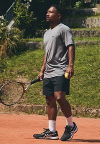 Camiseta atlética gris de manga corta con un pequeño logo, pantalones cortos negros, zapatillas de tenis negras, sosteniendo una raqueta de tenis y una pelota en una cancha de tierra batida.