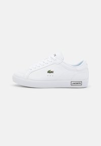 Baltas Lacoste kedus ar teksturētu ādas virsmu, plakaniem šņorēm, zaļu krokodila logotipu un melnu "LACOSTE" birku uz papēža.