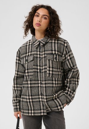 Jeune femme aux cheveux bouclés portant une veste à carreaux noire et beige boutonnée, un pantalon foncé et tenant un sac noir à bandoulière.