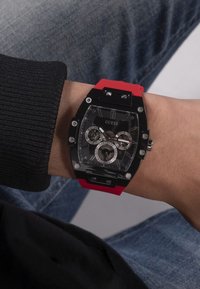 Black watch with a rectangular face, red silicone strap, silver hardware, and three subdials. Features a transparent case back.