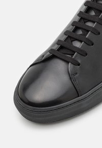 Svart läder sneaker med rund tå, svarta snörningar och texturerad gummisula, visad i närbild mot en vit bakgrund.