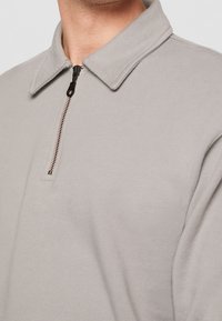 Suéter de color gris claro con cuello con cremallera, que presenta una textura de tela suave y una cremallera oscura en contraste. Diseño simple y minimalista.