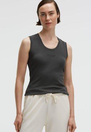 Woman with short hair wearing a dark gray sleeveless top and light beige drawstring pants, standing against a plain background.