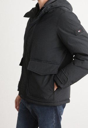 Homme portant une veste matelassée noire à capuche avec poches à rabat et un écusson Tommy Hilfiger sur le bras gauche, associée à un jean bleu.