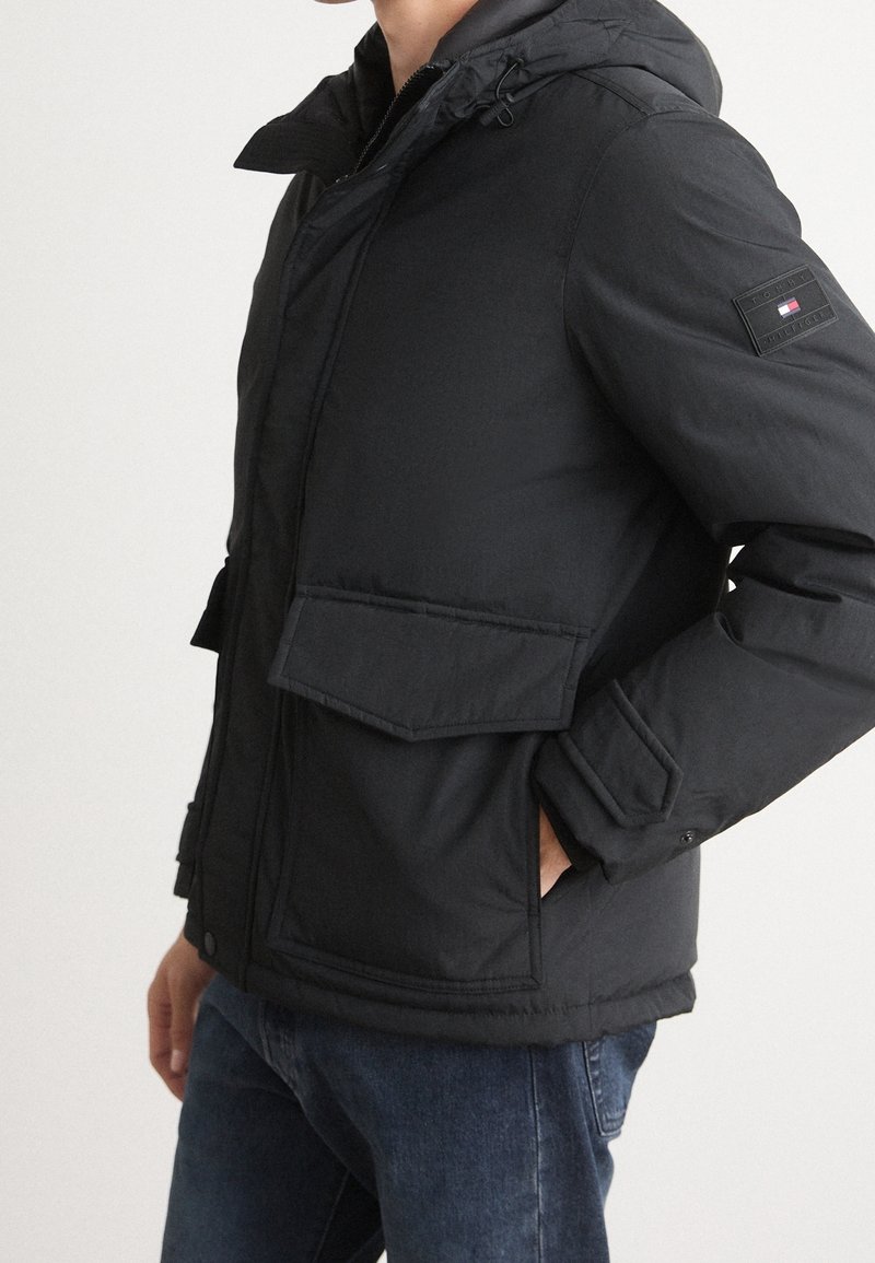 Homme portant une veste matelassée noire à capuche avec poches à rabat et un écusson Tommy Hilfiger sur le bras gauche, associée à un jean bleu.