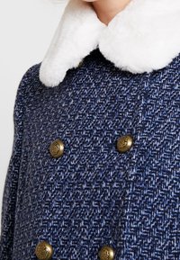 Dubbelknäppt marinblå kappa i strukturerat tyg, med vit fuskpälskrage och guldfärgade knappar. Har ett mönster i hundtandsdesign.