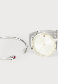Reloj de pulsera de malla plateada con una esfera redonda de color amarillo claro, acentos de cristal y logotipo de la marca. Acompañado de una pulsera minimalista de plata con un detalle de color.