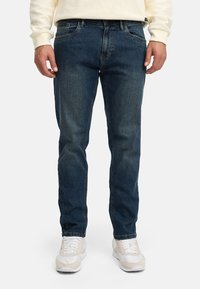 Man draagt blauwe rechtvallende jeans, een roomkleurige trui en witte sneakers, staand tegen een effen lichte achtergrond.