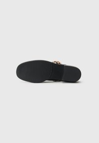 Black shoe sole with textured grip and gold buckle detail on upper, shown from bottom view against white background.