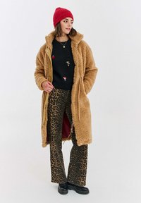 Žena v červené čepici, béžovém teddy kabátu, černém svetr s malými vyšitými nášivkami, kalhotách s leopardím potiskem a černých platformových botách.