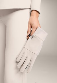 Jasnobeżowe rękawiczki wykonane z gładkiej tkaniny, z elastycznymi mankietami i trzema szwami na plecach. Logo widoczne na nadgarstku.