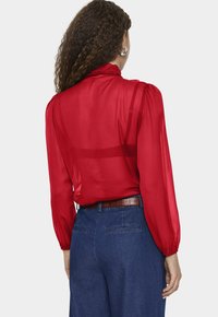 Piros, áttetsző blúz fodros nyakkal és hosszú, fodros ujjakkal, magas derekú farmernadrággal és barna texturált övvel párosítva.