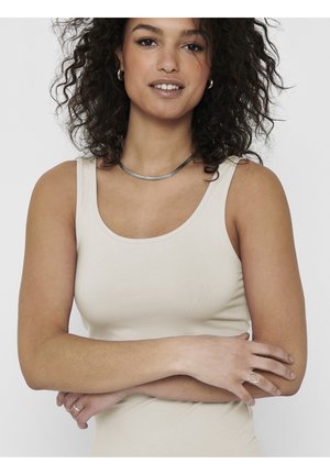 Woman with curly hair wearing a beige tank top, silver hoop earrings, a necklace, and crossed arms against a plain background.