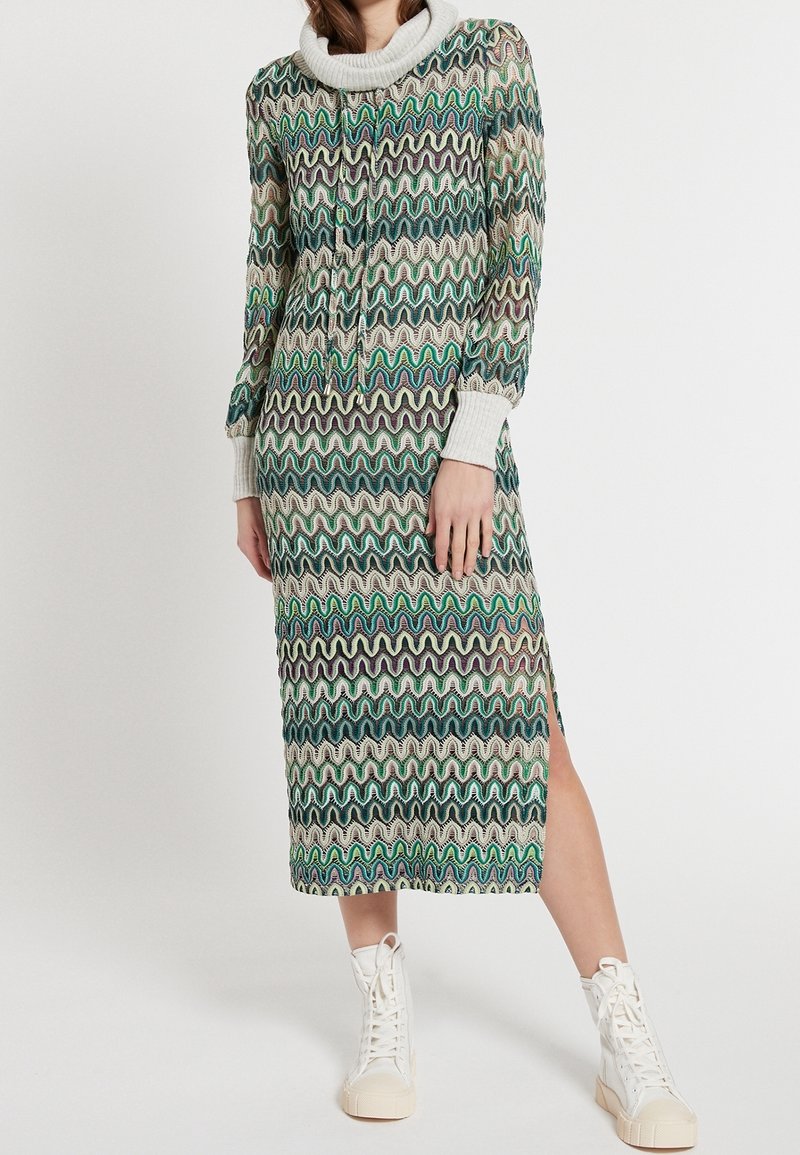 Długa dzianinowa sukienka z falistym wzorem w kolorach zielonym, fioletowym i kremowym. Ma kołnierz typu komin i ściągacze na rękawach. Boczne rozcięcie, zestawiona z białymi trampkami.