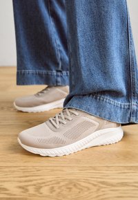Zapatillas deportivas de color beige claro con parte superior de malla, detalles texturizados y suela de goma blanca, combinadas con pantalones de mezclilla anchos de color azul sobre un suelo de madera.