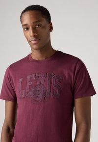 Bordeauxfarbenes Baumwoll-T-Shirt mit einer strukturierten Levi's-Logo-Grafik auf der Front. Kurze Ärmel und klassischer Rundhalsausschnitt.