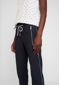 Närbild av en person som bär marinblå texturerade byxor med en framficka med dragkedja och vit kant, tillsammans med en vit mönstrad t-shirt.