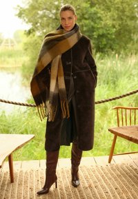 Barna gyapjú kabát nagy gombokkal, földszínekkel csíkos sállal párosítva. Hosszú barna bőr csizmák hegyes orral. Texturált háttér.
